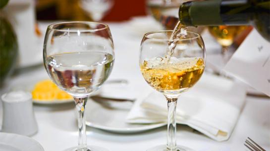 Sur une table un verre d'eau et un verre de vin blanc. Le verre de vin est en train d'être rempli.