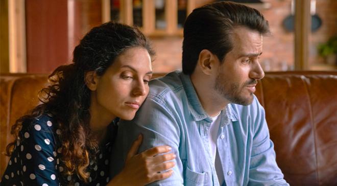 Un homme assis sur un canapé regarde dans le vague. Une femme assise à côté de lui le tient par l'épaule.